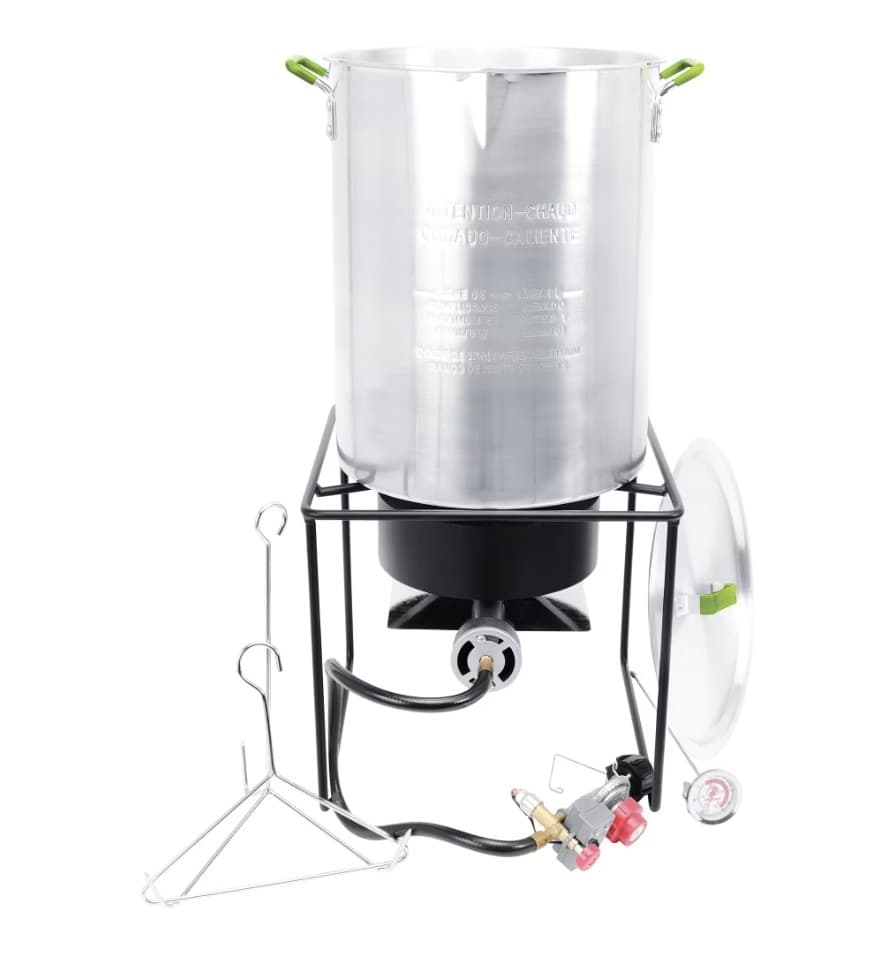 A tall aluminum pot with green handles on a black welded burner stand, with its lid and a dial thermometer leaning at the right, a wire turkey rack and lifting hook at the left, and the propane hose and regulator in front.
