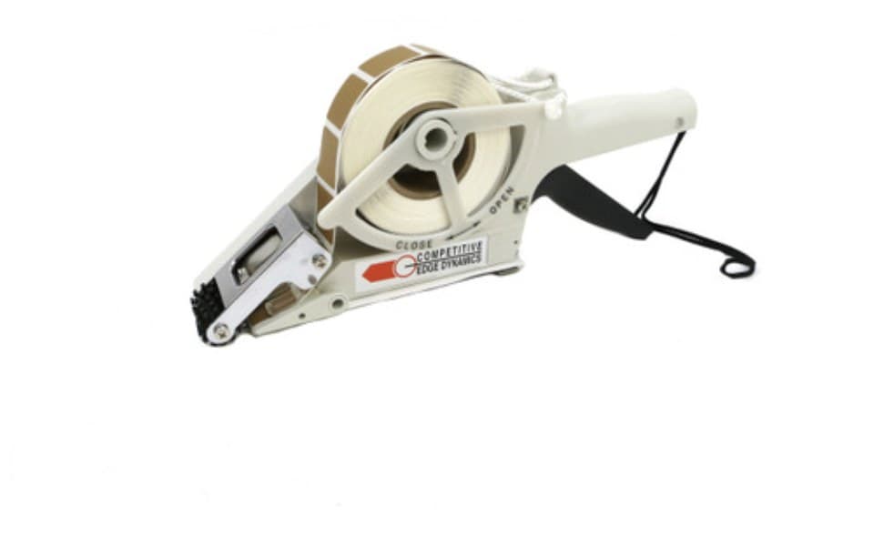 A CED Deluxe Quick Patch paster tape gun in cream and grey, lying at an angle: a roll of tan pasters mounted on the spool at the left, the applicator head below it and a pistol grip with a wrist lanyard at the right.