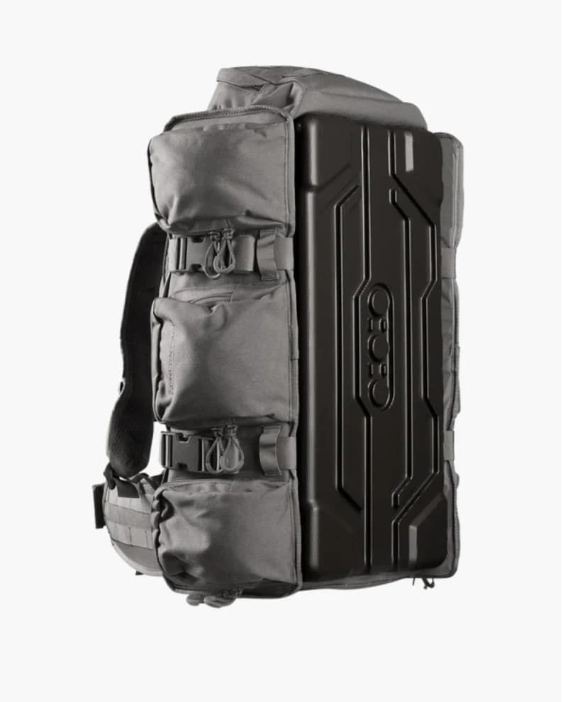 An Eberlestock UpRanger pack in gray, standing upright seen from the side: three zippered grey pockets stacked down the near face, the black moulded polycarbonate outer panel behind them, and the shoulder harness at the left.