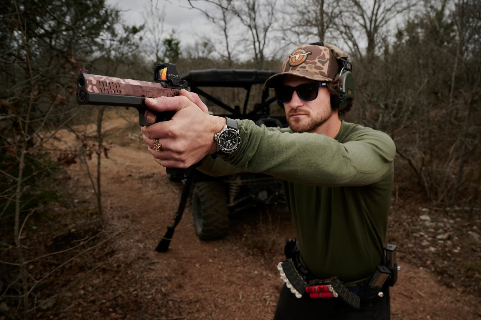 Riley at full extension with the Hayes Custom 2011 — ghosted rose PVD slide, Vortex Defender-XL red dot, black frame and magwell — on a ranch road with a side-by-side behind him.