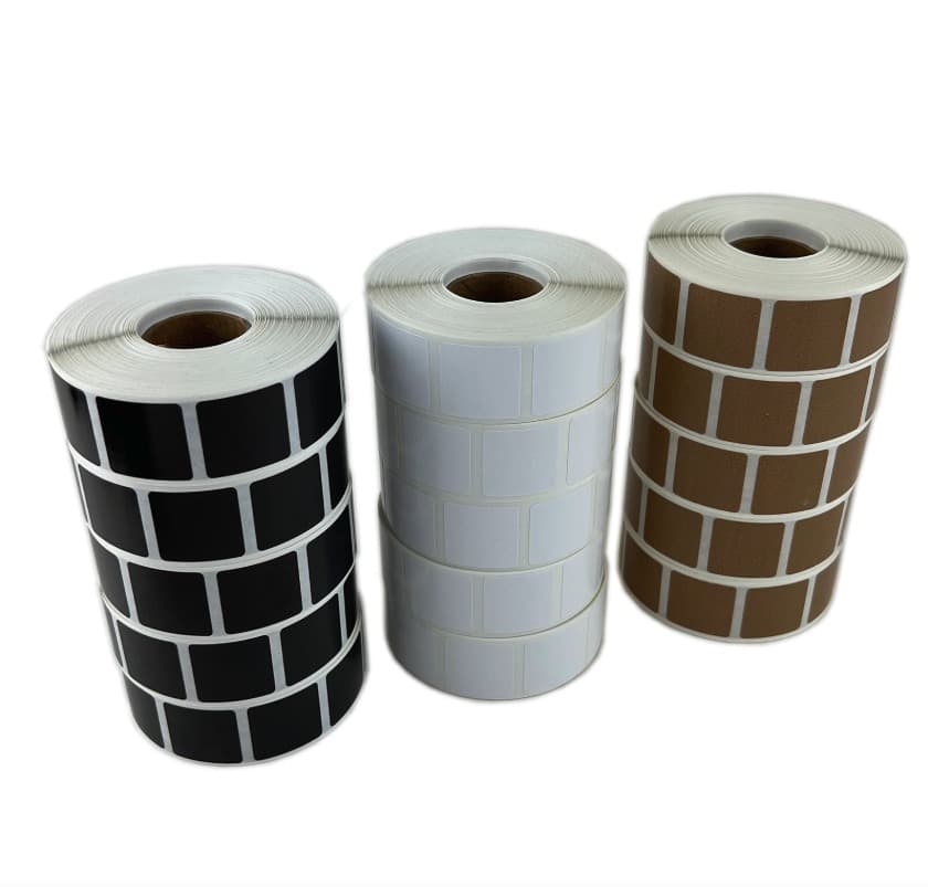 Three rolls of square target pasters standing on end, black, white and tan, each roll five pasters wide on a white backing.