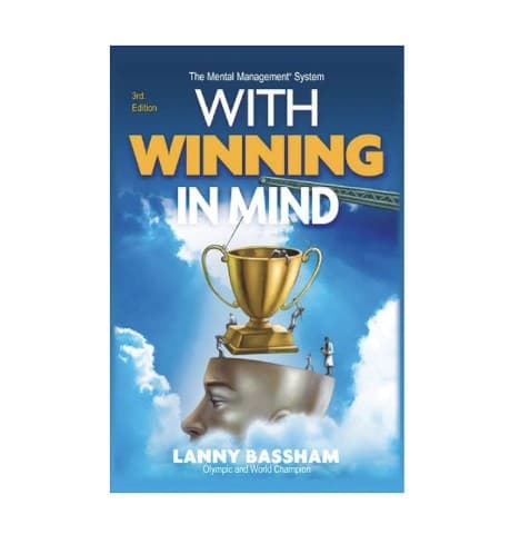 The cover of With Winning in Mind, 3rd Edition, by Lanny Bassham: a gold trophy being lowered by crane onto an open sculpted head against blue sky and clouds.