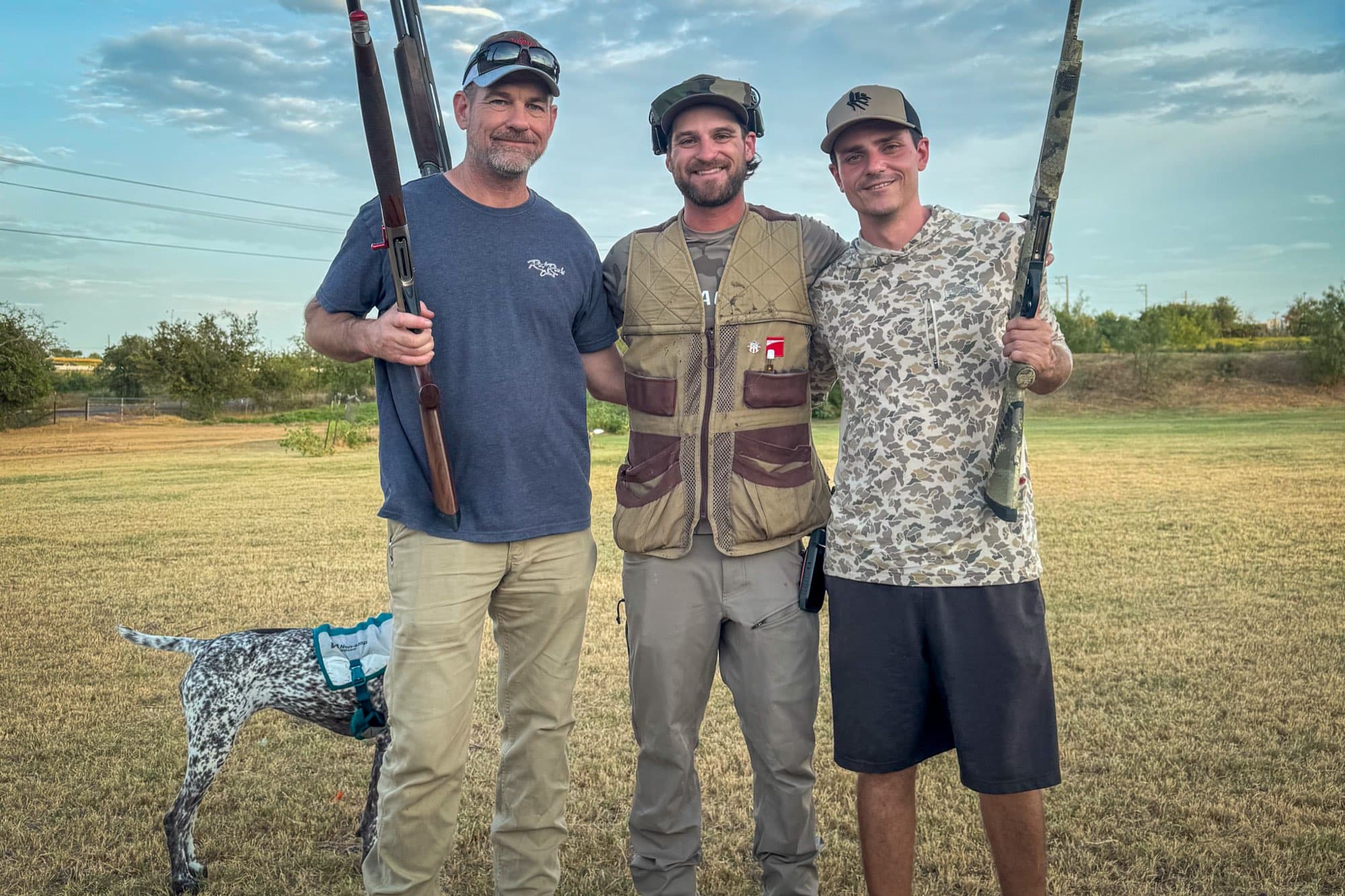 Three of the Hill Country Hitters standing in a Texas field at golden hour after a dove hunt, shotguns over their shoulders and a German shorthair at heel.